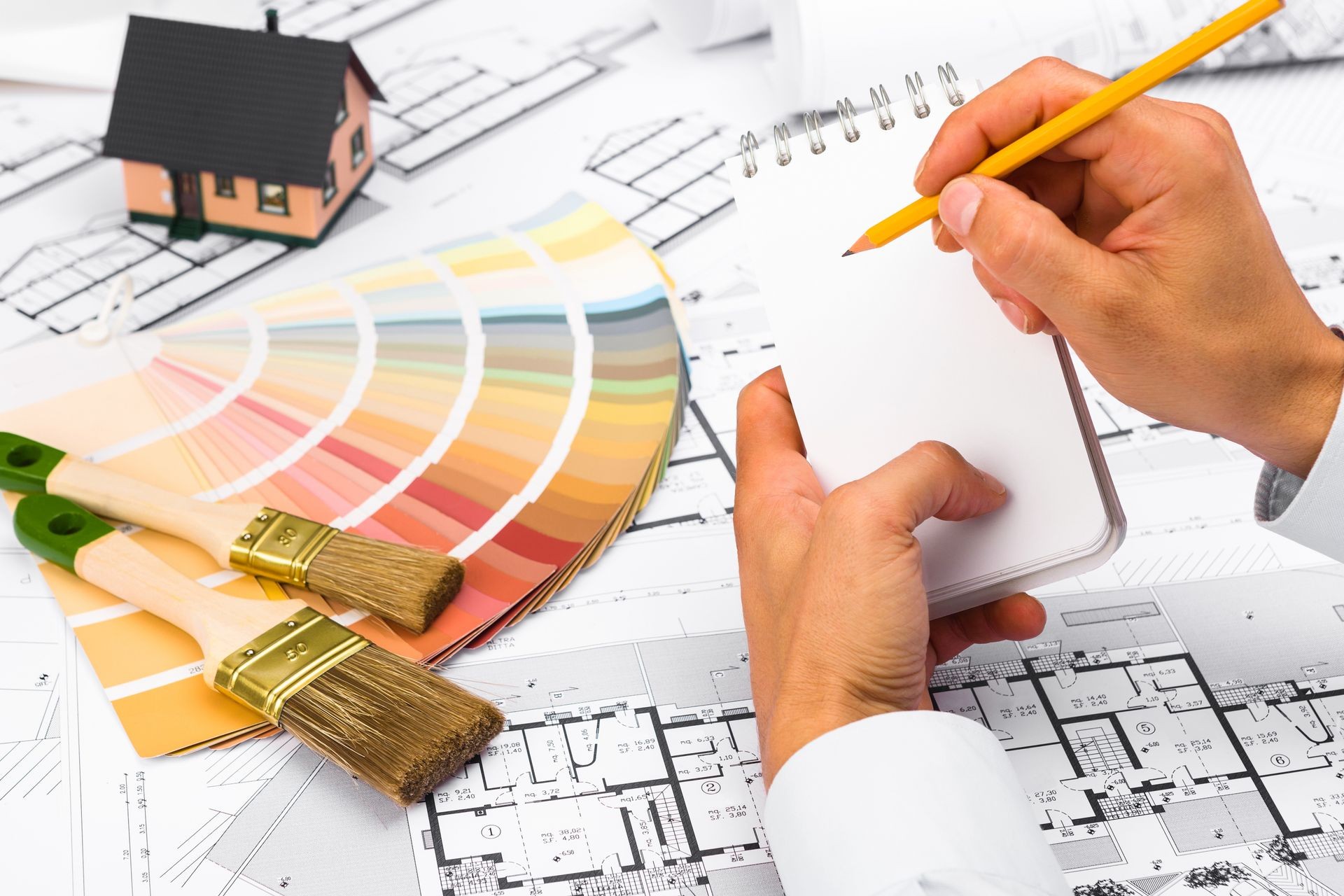 Male Hands writing on notebook; Construction plans with Color Palette and Miniature House on blueprints; Business and Construction Industry Concept
